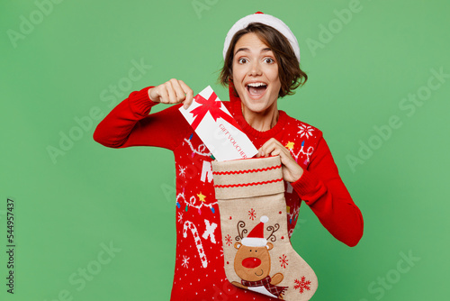 Merry fun young woman wear xmas sweater Santa hat posing hold stocking sock gift certificate coupon voucher card for store isolated on plain pastel light green background. Happy New Year 2023 concept.