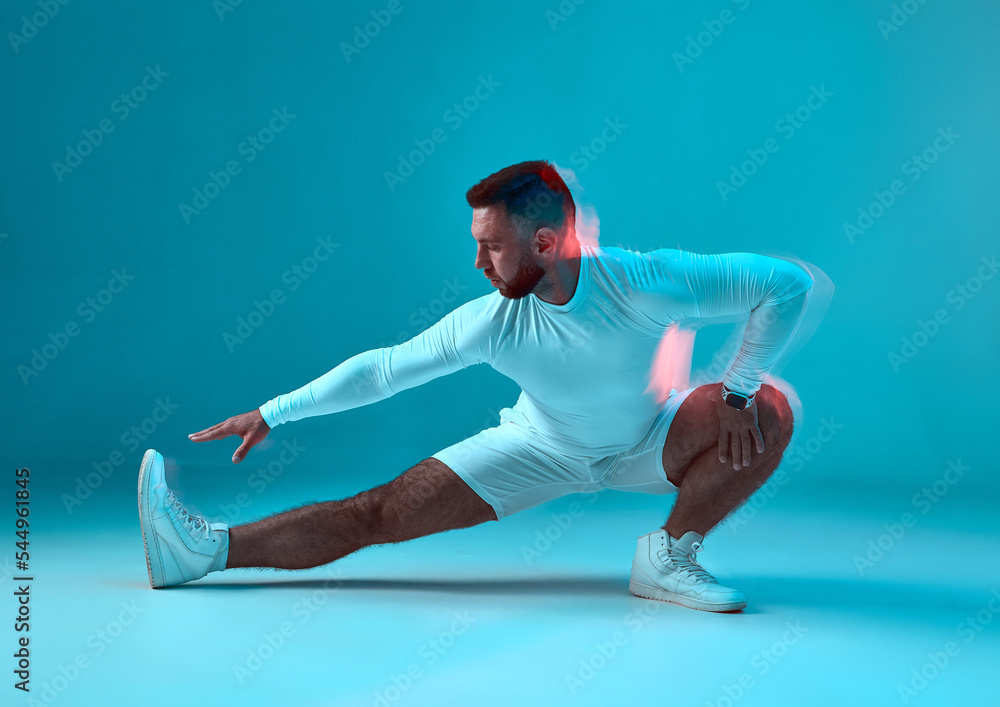 Cool sportive guy stretching leg on floor on blue studio background. Sport club advertisement. Gym, athletic training Cool sportive guy stretching leg on floor on blue studio background. Sport club advertisement. Gym, athletic training
