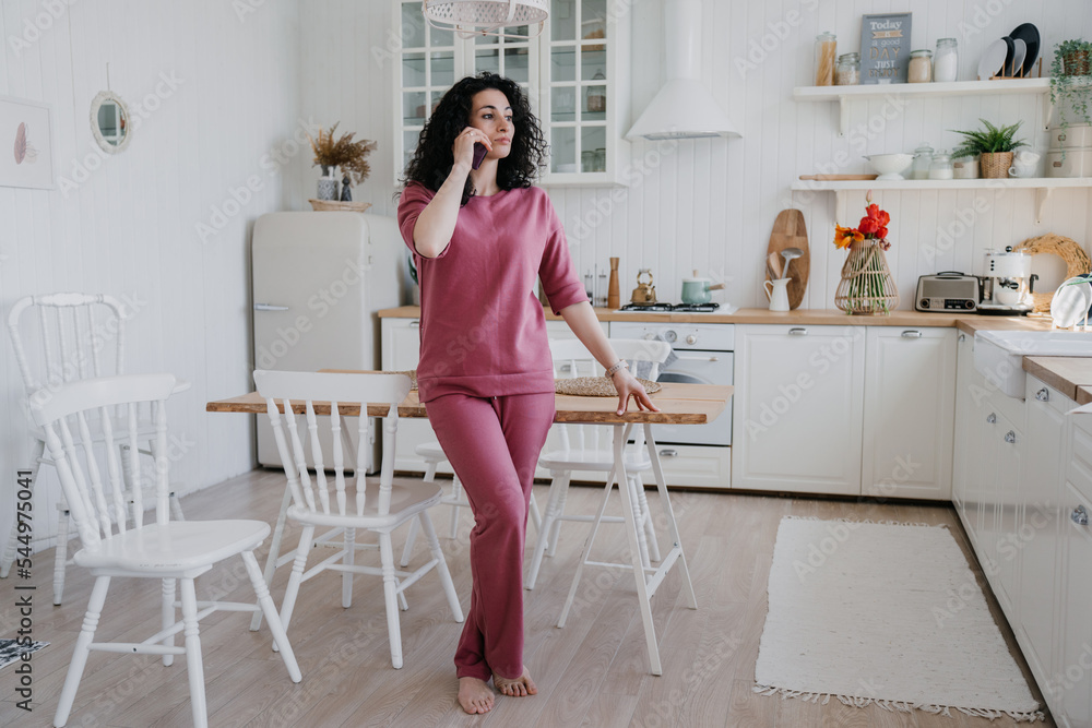 Young curly Italian woman talking by phone in lilac sweatshirt looking aside confidently. Caucasian pretty housewife at kitchen talking using smartphone. Serious arabic woman getting audio message.