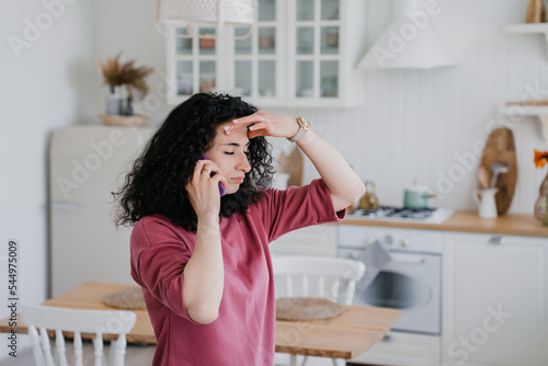 Young curly Italian woman talking by phone in lilac sweatshirt looking aside confidently. Caucasian pretty housewife at kitchen talking using smartphone. Serious arabic woman getting audio message.