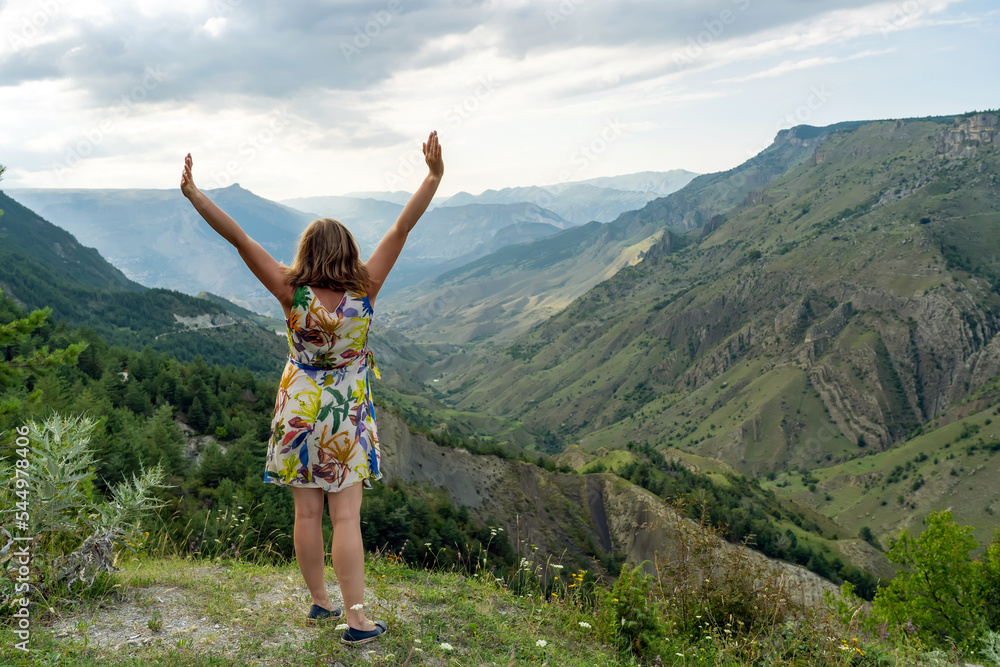 Fototapeta premium Young happy woman traveler in dress with outstretched arms standing on grassy hillside in summer mountains enjoying moment