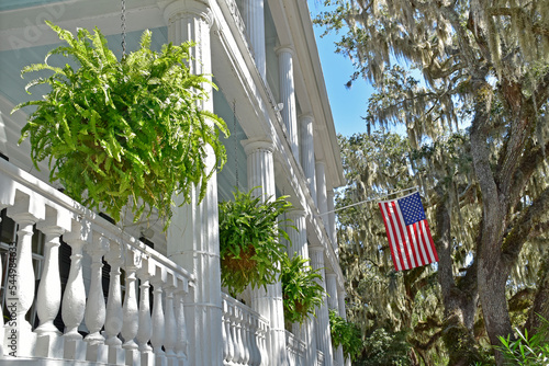 The columns and railings of an old southern home are graced with baskets of ferns and the American flag.