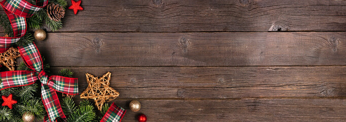 Christmas corner border of ornaments, branches and red and green plaid bows and ribbon. Top down view on a rustic wood banner background.