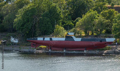 Helsinki, Finland - July 20, 2022: Suomenlinna Fortress from the sea. Small marroon-black-white submarine vessel put on blocks on shoreline with green foliage in back