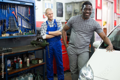Portrait of satisfied male client with mechanician standing near her car after repairing in auto workshop