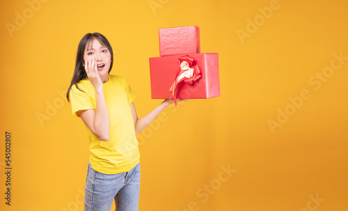 Cheerful excited asian woman holding two red and gold gift box in own hand while standing over isolated yellow background. Surprise greeting anniversary birthday or celebration Christmas and new year.
