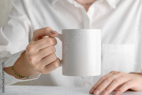 Female hand holding white mug mockup with blank copy space for your advertising text message or promotional content. Girl in white shirt holding white porcelain coffee mug mock up