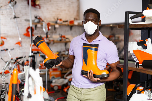 African-american man in face-mask choosing boots in gardening tools store.