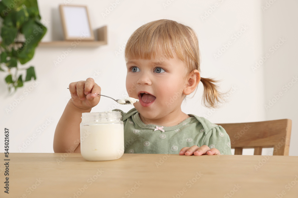 Cute little child eating tasty yogurt with spoon at wooden table indoors Cute little child eating tasty yogurt with spoon at wooden table indoors