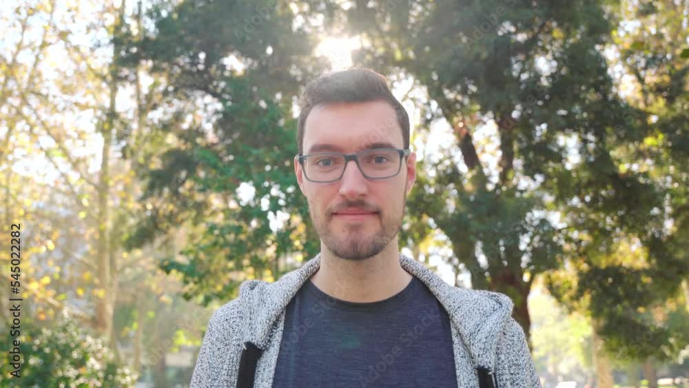 Young man glasses smiles sympathetically. Zoom in city park nature outdoor background sunflare. Modern caucasian male in twenties, beautiful portrait Young man glasses smiles sympathetically. Zoom in city park nature outdoor background sunflare. Modern caucasian male in twenties, beautiful portrait