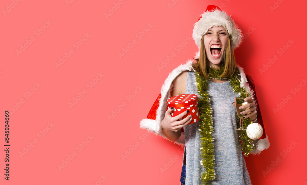 Funny winkingh woman with christmas ball and champagne. Pretty lady celebrate winter holidays hold christmas balls and champagne on red isolated background.