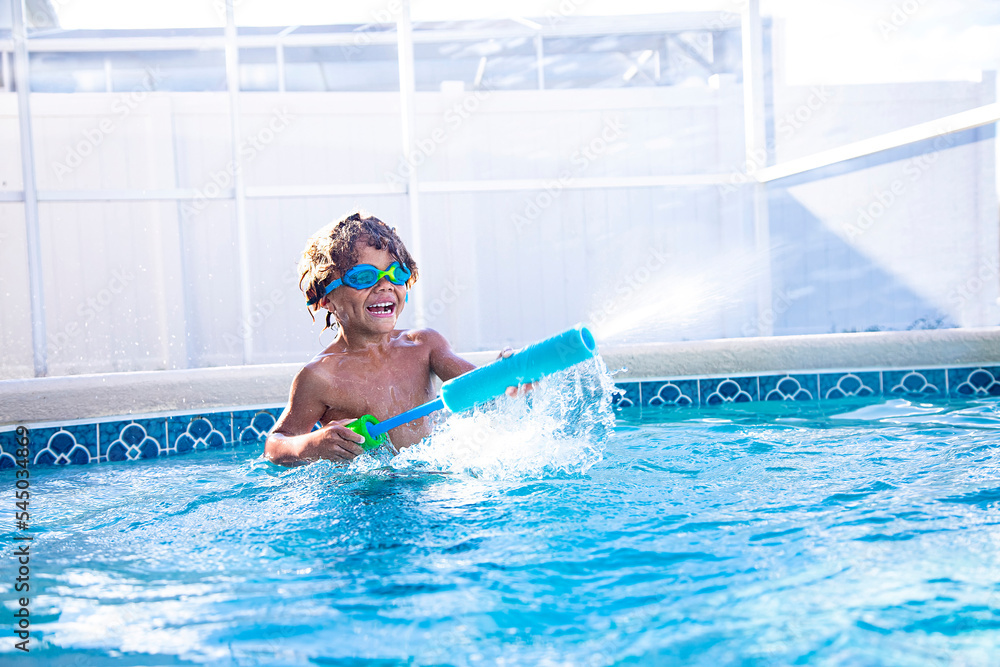 Cut African American little boy shooting a water gun while playing with friends in a backyard swimming pool. Laughing as he gets his friends wet playing games in the pool wearing goggles