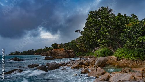 Evening on a tropical island. Boulders lie near the shore of the turquoise ocean. Waves are foaming on the rocks. Lush tropical vegetation against the sky and clouds. Seychelles. Mahe