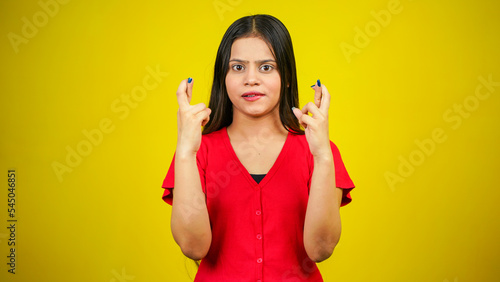 Portrait of a young girl with long hair standing over yellow background, holding fingers crossed for good luck