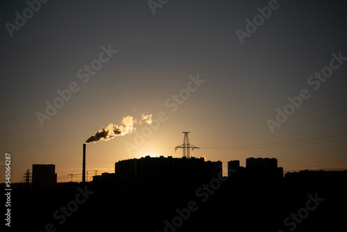 A dark silhouette of city buildings and a chimney at sunset. Industrial urban landscape.