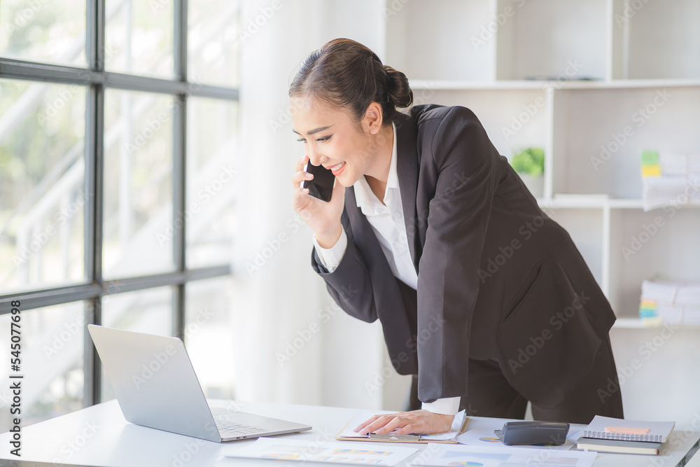 Young beautiful asian woman using smartphone and working with laptop while sitting at office desk, working from home concept.