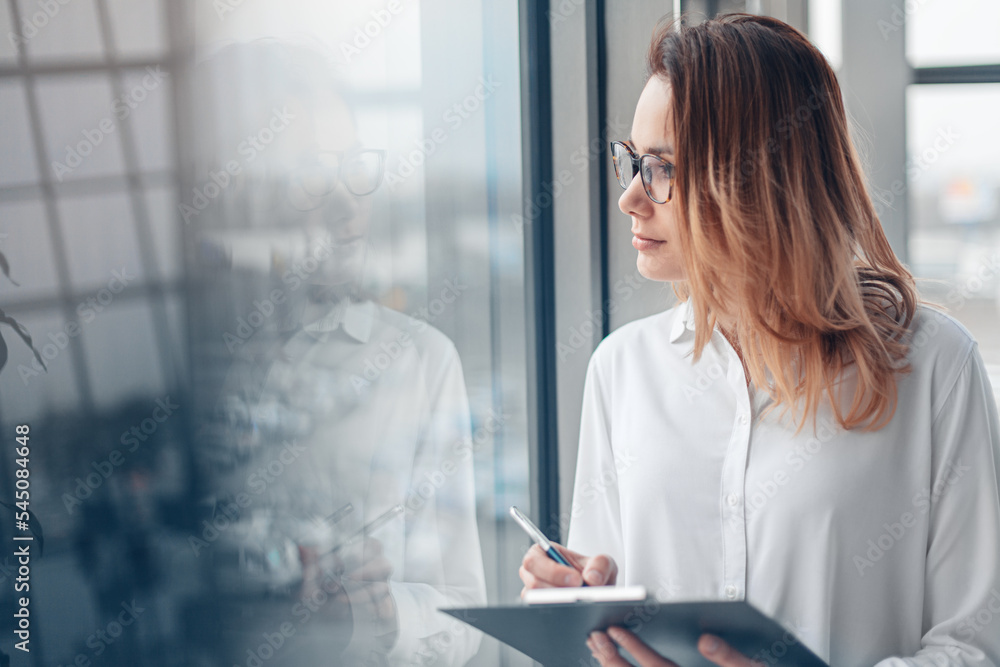Young woman holding folder of documents and standing at the panoramic window of a modern office.