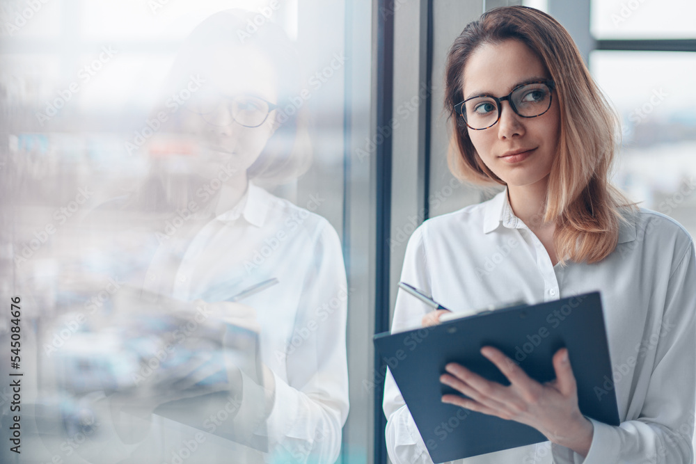 Young woman standing at the panoramic window of a modern office with a folder of documents and a pen.