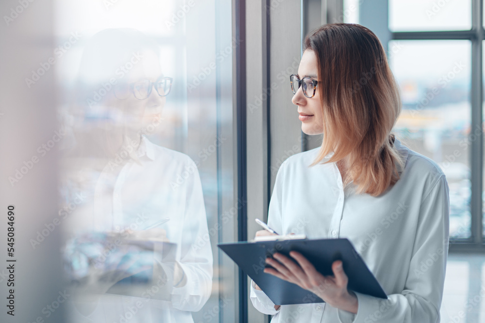Young woman looking in the panoramic window of a modern office with a folder of documents and a pen. Young woman looking in the panoramic window of a modern office with a folder of documents and a pen.