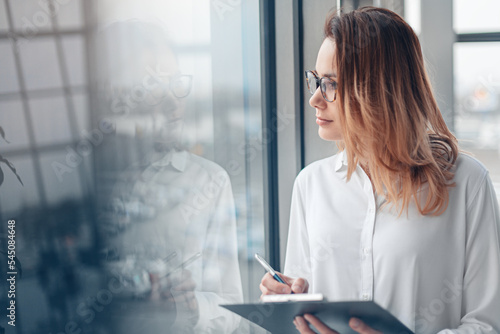 Young woman holding folder of documents and standing at the panoramic window of a modern office.