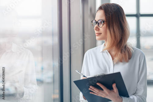Obraz na płótnie Young business woman holding folder of documents and standing at the panoramic window of a modern office. w salonie