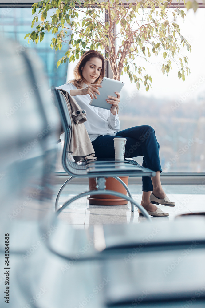 Young traveling woman using a tablet and waiting for a flight in the departure hall at the airport.