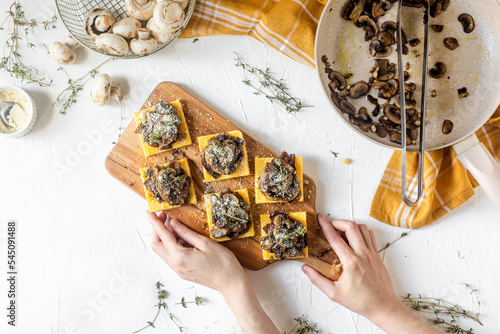 Tasty vegan polenta bites with cheese and grilled mushrooms on a white table with female hands holding the serving board, top view