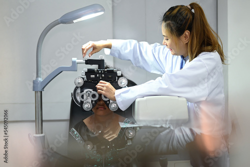 Fototapeta Naklejka Na Ścianę i Meble -  Young female ophthalmologist testing vision or sight of patient with optical phoropter in modern ophthalmology clinic