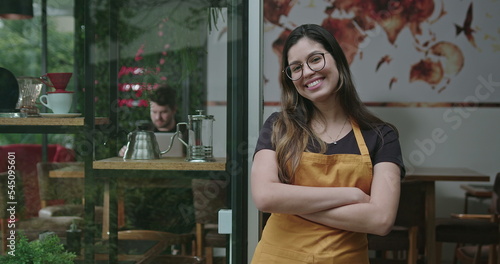 Happy female staff crossing arms smiling at camera in front of coffee shop store wearing yellow apron. A hispanic latin adult girl barista employee of small business restaurant place