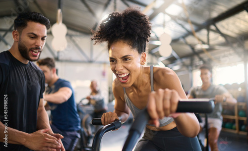 Fototapeta Naklejka Na Ścianę i Meble -  Gym, fitness and woman on bike with personal trainer for motivation, support and power workout, cheering and clapping hands. Coach, black woman and cycling exercise at sport center, sweat and energy