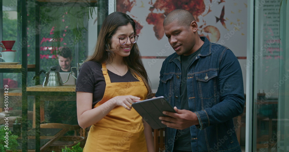An African American manager of small business holding tablet speaking with female employee at coffee shop entrance. Portrait of two diverse happy staff of restaurant cafe place