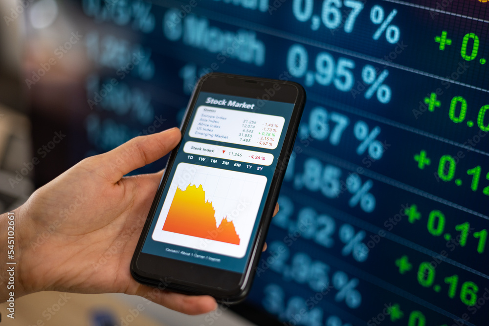 Woman's hand holds a cellphone with falling stock index chart on the display. Computer monitor with financial figures in the background. Stock market crash, office, banking trading and business.