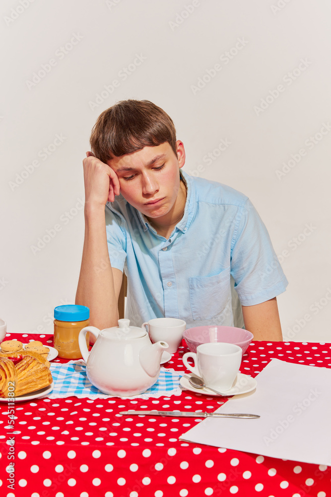Portrait of young boy in blue shirt sitting at the table and having breakfast isolated on grey background. Bored Portrait of young boy in blue shirt sitting at the table and having breakfast isolated on grey background. Bored