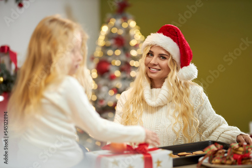 Front view of blonde woman wearing christmas hat, looking at camera, smiling, baking cookies with daughter. Family having photoshoot indoors, posing. Concept of winter holiday. © serhiibobyk