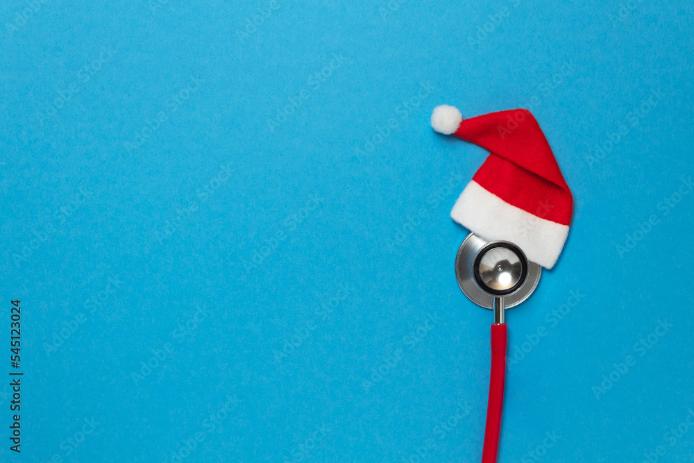 Medical red stethoscope with christmas hat on blue background. Christmas and New Year concept. Creative medical winter greeting card. Top view, flat lay, copy space Medical red stethoscope with christmas hat on blue background. Christmas and New Year concept. Creative medical winter greeting card. Top view, flat lay, copy space