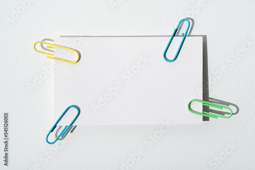 Close-up white sheet with copy space attached paper clips. Business card with colorful paper clips on white background. Sheet of paper for notes and clip. Sticker note with clip. Concept for notes