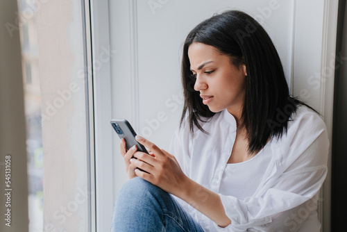 Perplexed young African American wonan in white shirt holds phone sitting on windowsill with upset facial expression reading message at home. Exhausted Brazilian girl received unpleasant news.