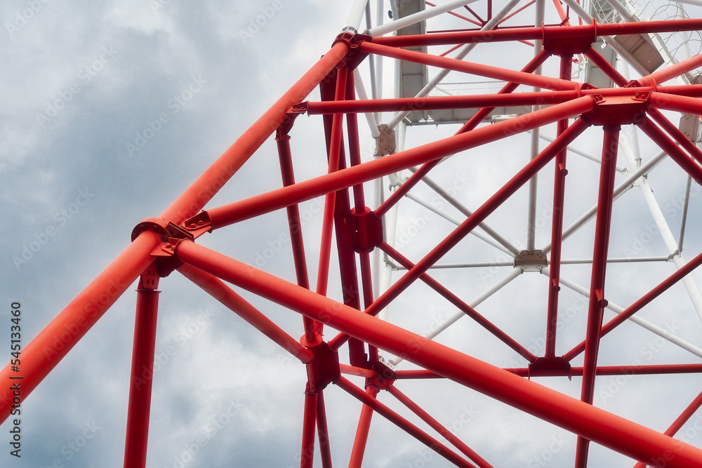 View of part of metal structures of communication tower against background of cloudy sky. Red metal pipes. Industrial background.