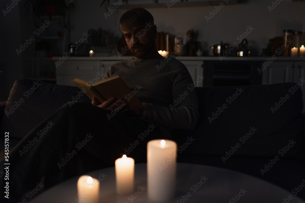 man sitting in kitchen during energy blackout and reading book near lit candles.