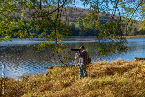 A male photographer using his monopod and long lens stands on the edge of a lake taking photos of the far shoreline at Hawkins Pond Park in Broome County in Upstate NY.
