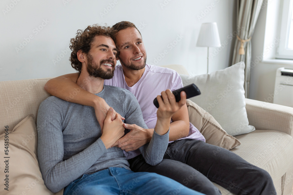 Young homosexual couple relaxing on sofa in the living room, embracing and enjoying while watching TV Young homosexual couple relaxing on sofa in the living room, embracing and enjoying while watching TV