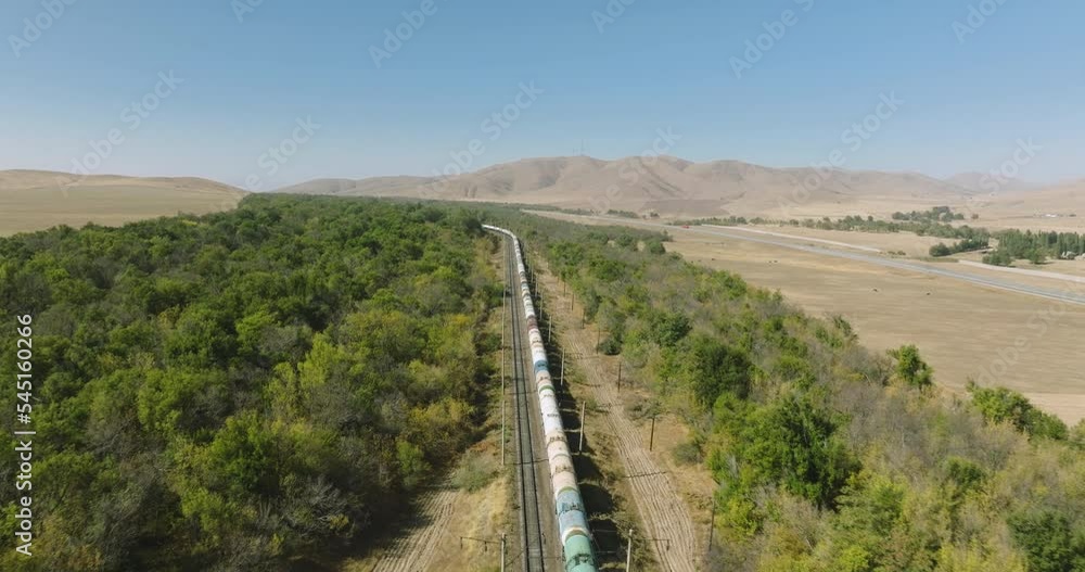 Freight long cargo train carries lorry carriages by arid hilly landscape. Railways in pampas. Aerial drone perspective view at summer sunset Freight long cargo train carries lorry carriages by arid hilly landscape. Railways in pampas. Aerial drone perspective view at summer sunset