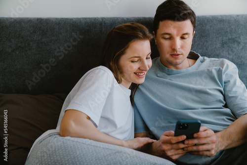 White young couple using mobile phone while resting on bed together