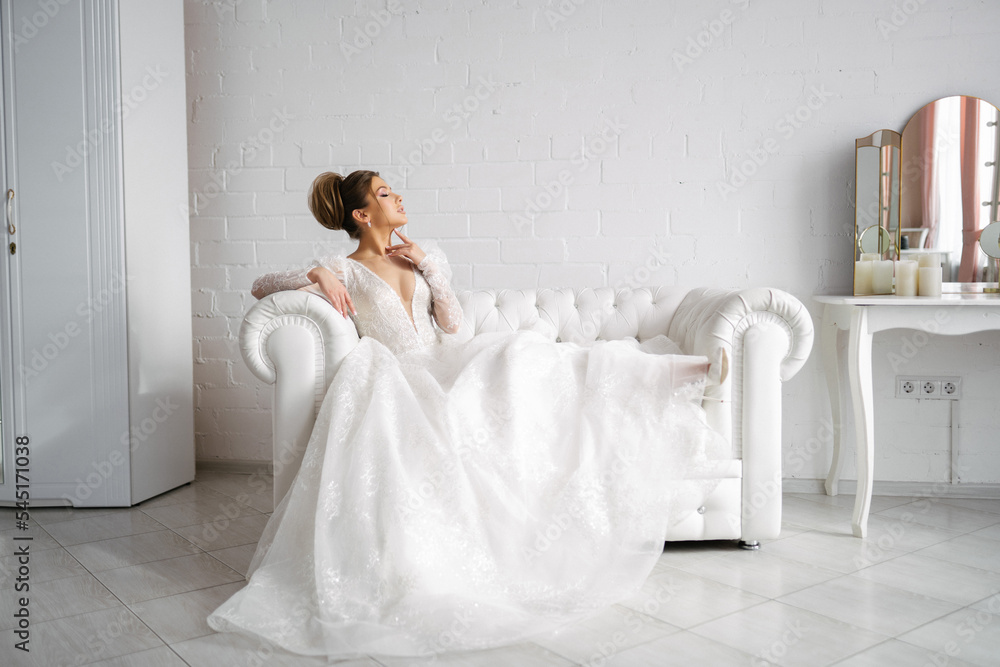 the bride in a wedding dress sits on the sofa spreading her train in a bright interior, gently touching her neck with her finger and enjoying the moment