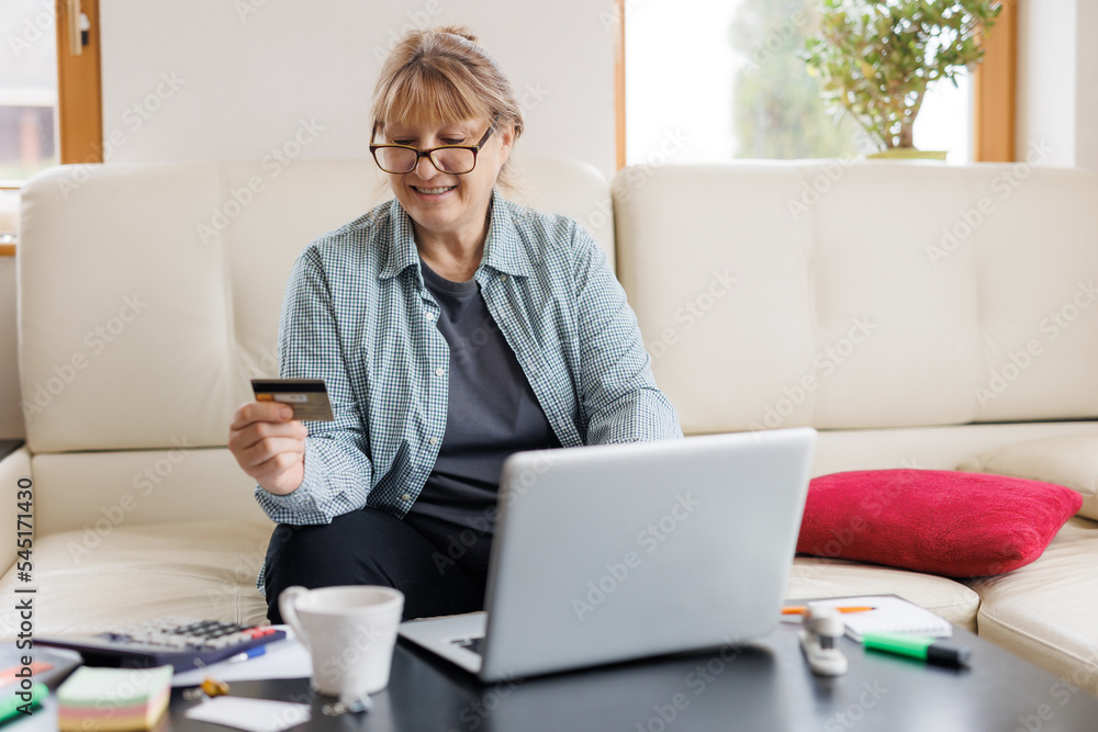 Happy mature woman grandmother using credit card , doing online shopping on laptop, buying new clothes, presents, paying domestic bills, pension at home Happy mature woman grandmother using credit card , doing online shopping on laptop, buying new clothes, presents, paying domestic bills, pension at home