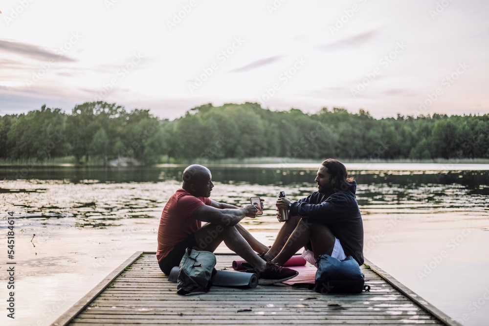 Man sharing smart phone with friend sitting on jetty at lake Man sharing smart phone with friend sitting on jetty at lake