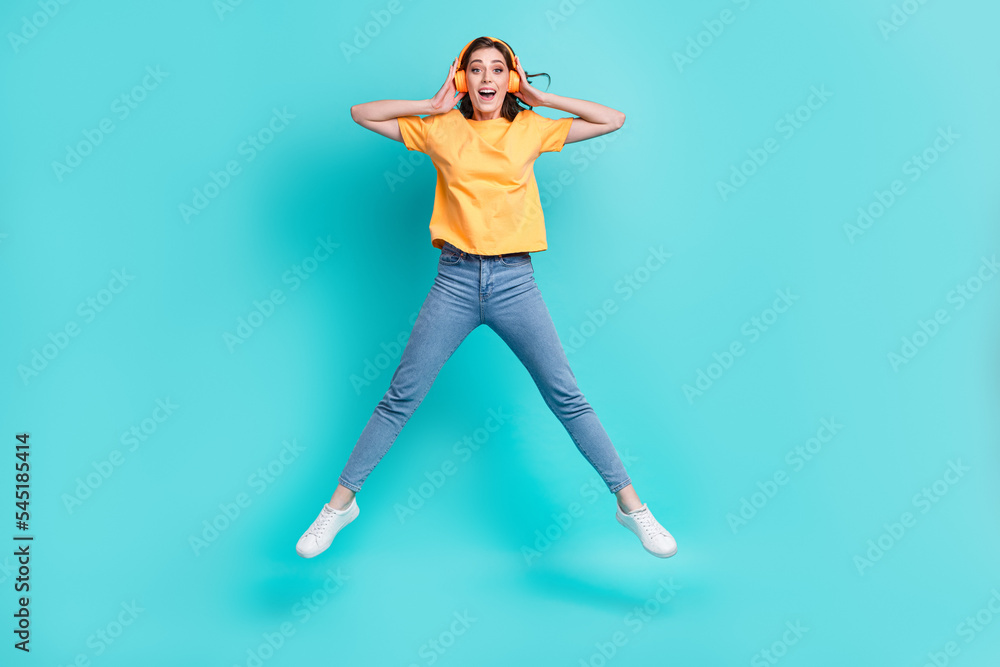 Full length photo of pretty impressed lady wear yellow t-shirt jumping high listening music isolated turquoise color background Full length photo of pretty impressed lady wear yellow t-shirt jumping high listening music isolated turquoise color background