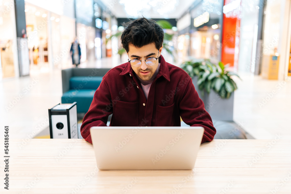 Portrait of focused bearded businessman in glasses sitting on sofa in shopping mall typing working on laptop. Front view of serious male freelancer using online application working remote, e-learning. Portrait of focused bearded businessman in glasses sitting on sofa in shopping mall typing working on laptop. Front view of serious male freelancer using online application working remote, e-learning.