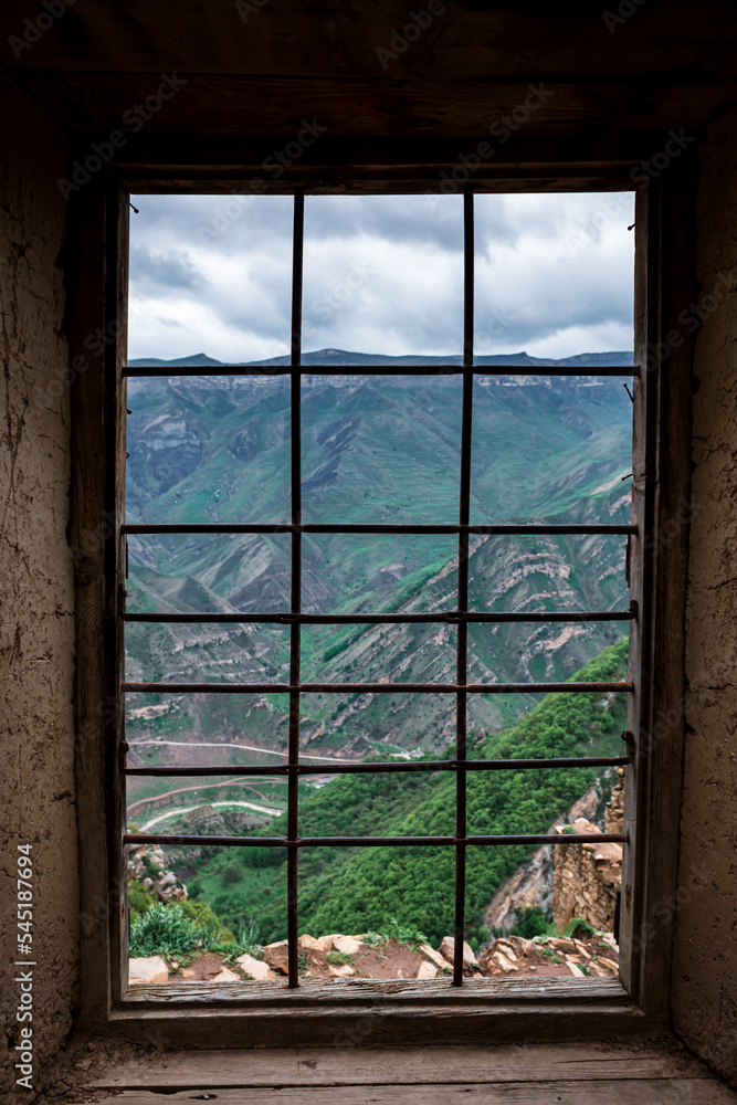 An ancient non-residential, stone city high in the Gamsutl mountains, against the backdrop of large, high mountains. View from the window with bars. Vertical Orientation An ancient non-residential, stone city high in the Gamsutl mountains, against the backdrop of large, high mountains. View from the window with bars. Vertical Orientation