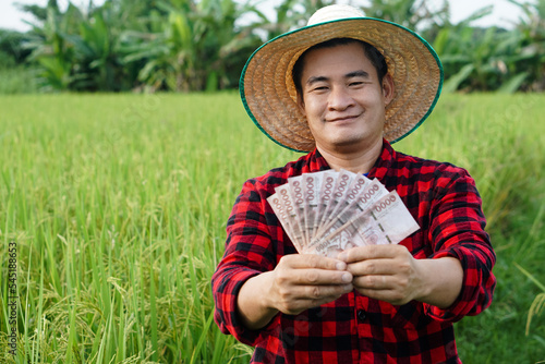 Asian farmer man is at paddy field, wears hat and red plaid shirt, hold Thai banknote money.  Concept : Farmer happy to get profit, income, agriculture supporting money.  